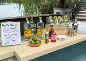 Poolside tequila bar setup on a lakeside patio with a margarita recipe sign, tequila and sparkling water bottles, a wooden bowl of limes, plastic glasses rimmed with lime wheels, cocktail shaker and jiggers.