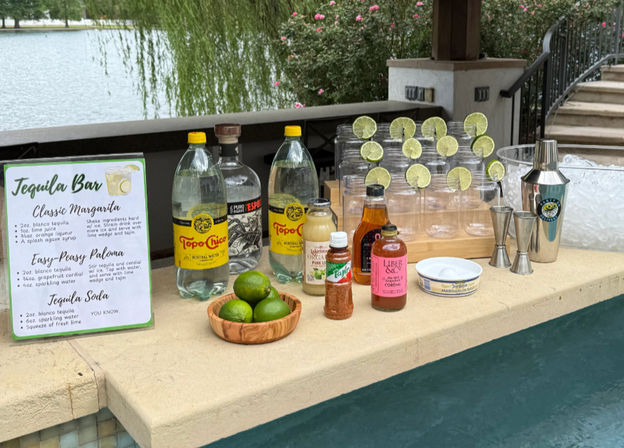 Poolside tequila bar setup on a lakeside patio with a margarita recipe sign, tequila and sparkling water bottles, a wooden bowl of limes, plastic glasses rimmed with lime wheels, cocktail shaker and jiggers.
