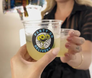 Close-up of two hands clinking clear plastic cups with pale yellow cocktails at an indoor celebration; one cup shows a round sticker with a cocktail glass and floral design, host blurred in the background.