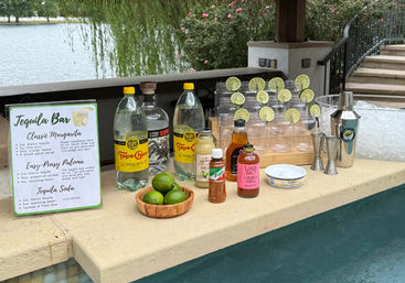 Lakeside poolside tequila bar setup with margarita recipe sign, bottles and mixers, wooden bowl of limes, stacked plastic cups garnished with lime slices, cocktail shaker, jiggers and ice bucket — outdoor margarita station.