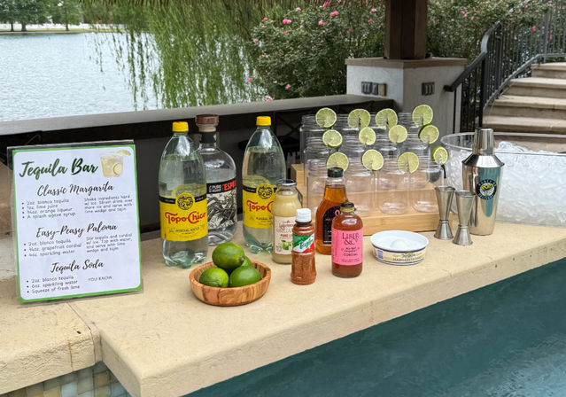 Lakeside poolside tequila bar setup with margarita recipe sign, bottles and mixers, wooden bowl of limes, stacked plastic cups garnished with lime slices, cocktail shaker, jiggers and ice bucket — outdoor margarita station.