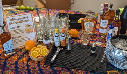 Hill-Country Old-Fashioned cocktail station on a colorful paisley tablecloth: stacked rocks glasses, bourbon bottles, bitters, jigger and bar tools, sliced and whole oranges, toothpicks, mirrored ice bucket and printed recipe sign.