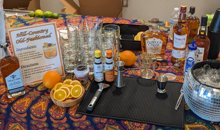 Hill-Country Old-Fashioned cocktail station on a colorful paisley tablecloth: stacked rocks glasses, bourbon bottles, bitters, jigger and bar tools, sliced and whole oranges, toothpicks, mirrored ice bucket and printed recipe sign.