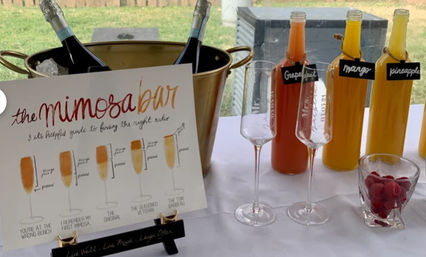 Backyard brunch mimosa bar with a gold ice bucket holding two champagne bottles, a handwritten mimosa bar sign showing drink styles, three labeled juice bottles (grapefruit, mango, pineapple), two champagne flutes and a small glass of raspberries on a white tablecloth.