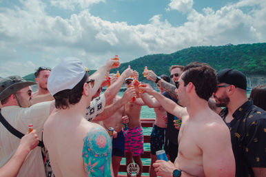 Group of friends on a summer lake boat, shirtless and in swim trunks, raising small bottled drinks in a celebratory toast with green wooded hills and a partly cloudy blue sky in the background.