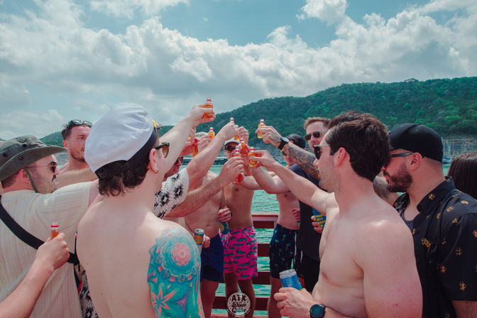 Group of friends on a summer lake boat, shirtless and in swim trunks, raising small bottled drinks in a celebratory toast with green wooded hills and a partly cloudy blue sky in the background.