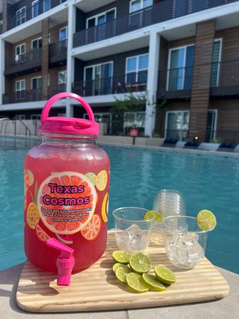 Poolside summer scene at a modern apartment complex: large pink fruit drink dispenser on a wooden board with two plastic cups of ice and lime slices, halved limes scattered, and a turquoise swimming pool in the background.
