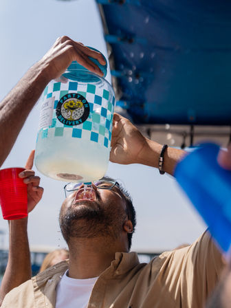 Outdoor summer party scene: friends hold a large blue-checkered jug and pour a cold drink into a man leaning back with mouth open, a red plastic cup nearby and bright daylight overhead.