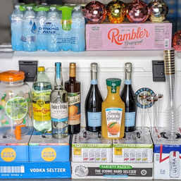 Party beverage display on a counter with bottled water and grapefruit sparkling packs, vodka and hard seltzer cases, tequila and prosecco bottles, orange juice and plastic champagne flutes — ready for a brunch or celebration