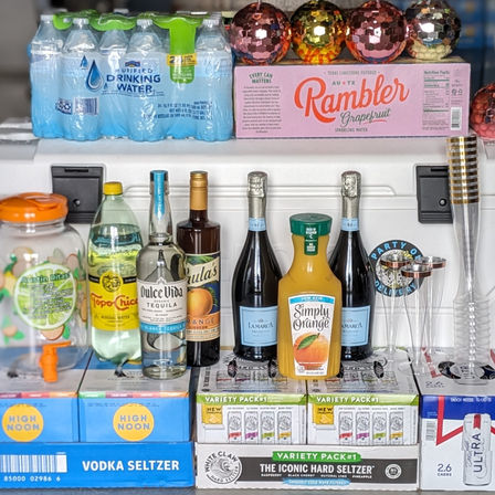 Party beverage display on a counter with bottled water and grapefruit sparkling packs, vodka and hard seltzer cases, tequila and prosecco bottles, orange juice and plastic champagne flutes — ready for a brunch or celebration