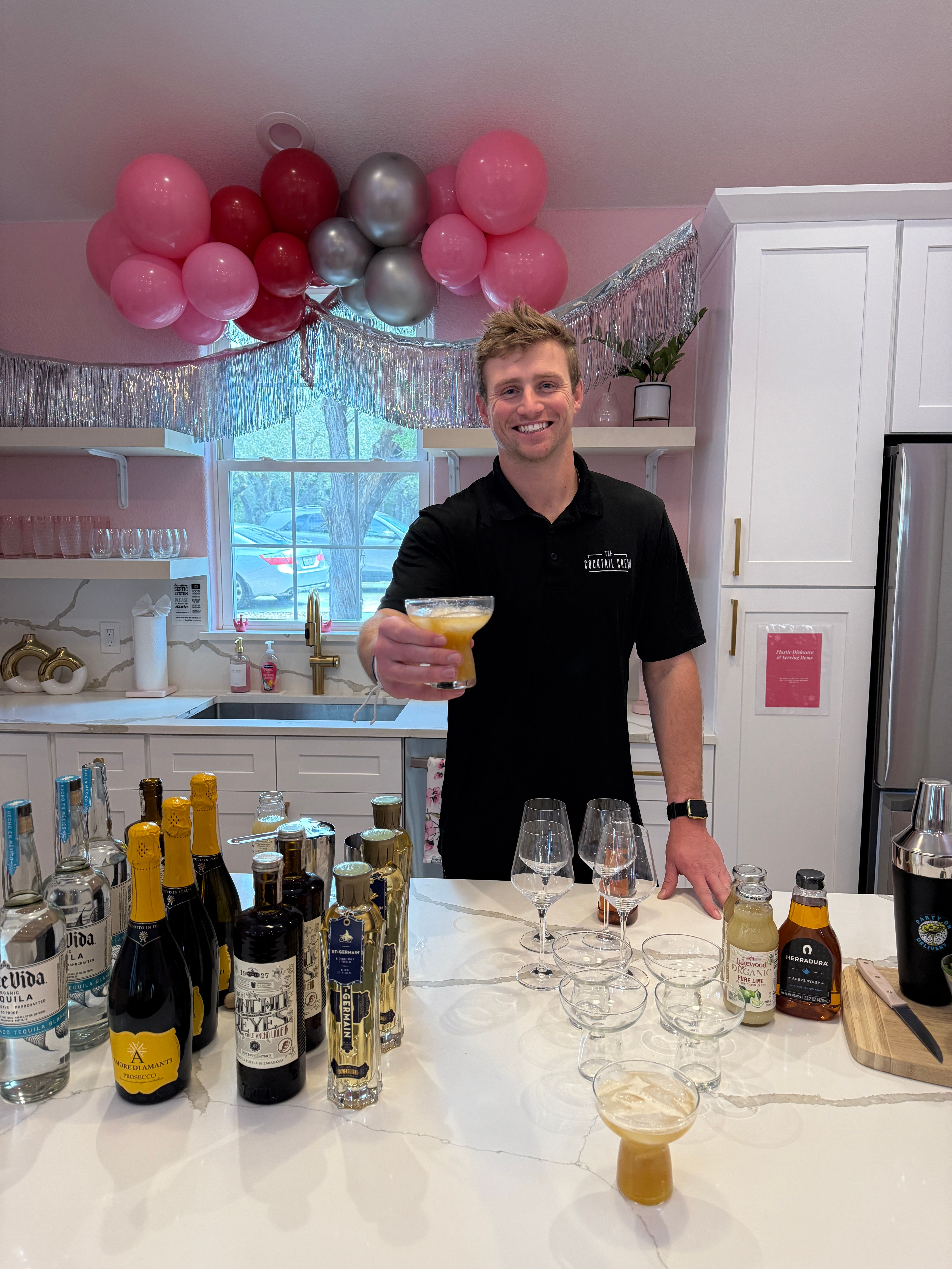 Smiling bartender offering a cocktail in a pink-decorated kitchen with pink and silver balloons, silver fringe, bottles and glassware on a marble island.
