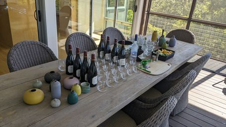 Sunlit outdoor wood dining table on a balcony deck with wicker chairs, set for a wine tasting — rows of wine bottles, empty tasting glasses, colorful ceramic vases, and a tray with lemon wedges and pitchers.