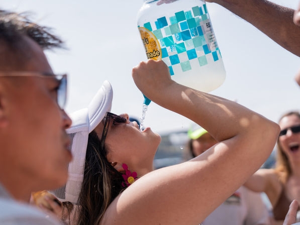 Woman in white cap and sunglasses chugs lemonade from a large blue-checkered jug at a sunny summer outdoor party as friends cheer