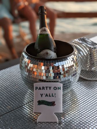 Champagne bottle chilling in a mirrored disco-ball ice bucket on a sparkly table with a “PARTY ON Y’ALL!” koozie, beach sunset party vibe