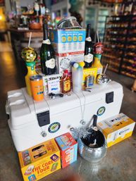 Party-ready liquor store display on a white cooler: champagne bottles, hard seltzer and beer packs, mixers, plastic cups, disco balls and festive drinkware