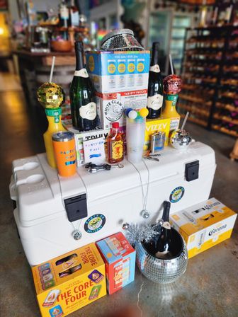 Party-ready liquor store display on a white cooler: champagne bottles, hard seltzer and beer packs, mixers, plastic cups, disco balls and festive drinkware