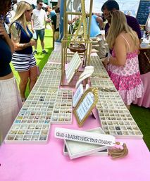 Outdoor craft fair jewelry stall with a pink tablecloth and rows of compartment trays filled with charms and beads as shoppers browse and pick pieces for custom bracelets.