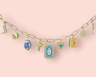 Close-up of a gold link charm bracelet on a pastel pink background featuring eclectic charms — cowboy boot, folded-leaf charm, blue floral bead, bull skull, blue hamsa hand plaque, heart with turquoise, tiny envelope locket, filigree disc and seahorse.