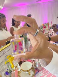 Hands holding a small perfume bottle in the foreground at a fragrance-blending workshop — pink-lit room, floral table display, attendees sampling scents.