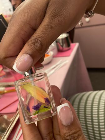 Close-up of a hand with glossy French-tip nails spraying a small glass perfume bottle filled with dried flower petals on a pink table in a manicure/beauty setting