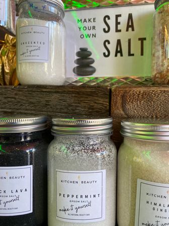 Rustic shop display of glass jars filled with bath salts (peppermint, black lava, Himalayan) on a wooden shelf under a 'Sea Salt' sign