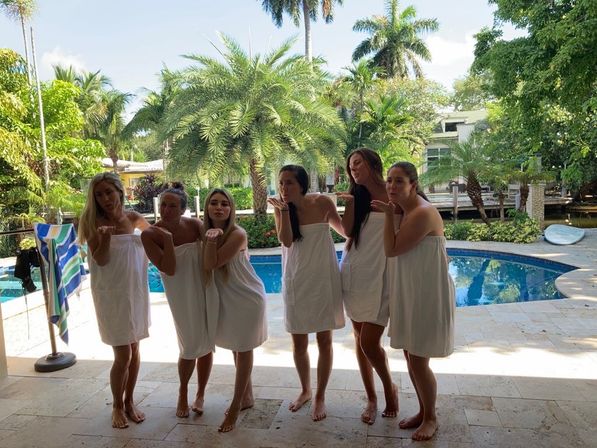 Six women in white towels standing barefoot on a sunny patio, playfully blowing kisses by a blue backyard pool framed by palm trees and lush tropical landscaping.
