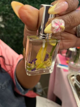Close-up of hand with yellow stiletto nails and pink-white 3D flower nail art holding a glass perfume spray bottle filled with yellow and purple floral petals