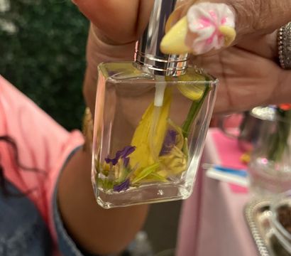 Close-up of hand with yellow stiletto nails and pink-white 3D flower nail art holding a glass perfume spray bottle filled with yellow and purple floral petals