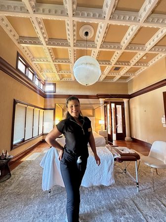 Smiling massage therapist posing in a sunlit luxury spa suite with ornate coffered ceiling, globe pendant light and massage table