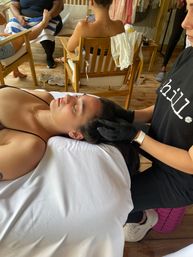 Relaxed woman lying on a white-sheeted table receiving a scalp/head massage from a gloved therapist in a wooden-deck spa seating area — peaceful wellness treatment.