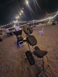 Nighttime beachfront event scene with warm string lights overhead, sandy ground, scattered folding tables and chairs, and a black portable massage chair in the foreground.