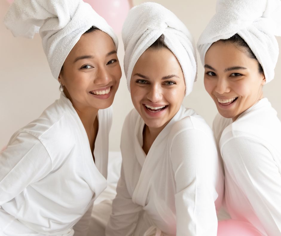 Three women in white robes with towel turbans smiling together during a bright spa-day pampering session