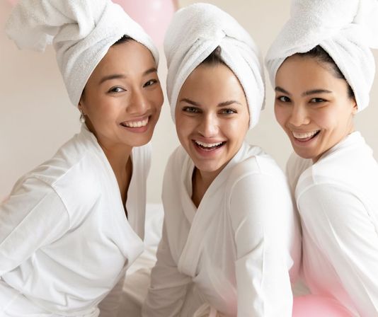 Three women in white robes with towel turbans smiling together during a bright spa-day pampering session