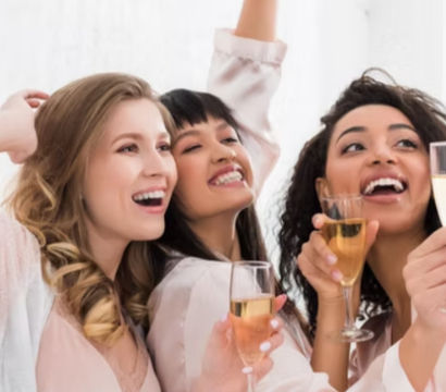 Three smiling women in pastel robes toasting with champagne glasses in a bright indoor setting, joyful bachelorette-style celebration