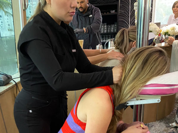 Massage therapist in black uniform giving a seated neck and shoulder massage to a woman leaning on a pink padded face cradle in an indoor lounge or event space with a bar counter, glassware and flowers in the background.