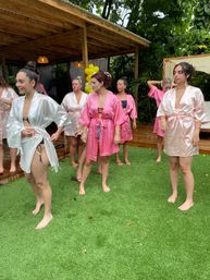 Group of women in pink and white satin robes on artificial lawn by a wooden cabana with yellow balloons and tropical greenery — playful outdoor bridal party getting ready.