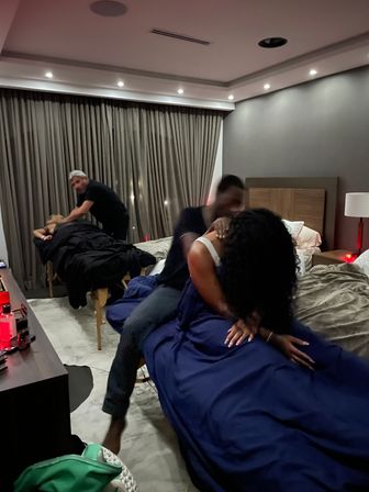 Dimly lit modern bedroom suite with two people receiving in-room massages—therapists working beside navy-covered beds, floor-to-ceiling curtains and warm bedside lighting.