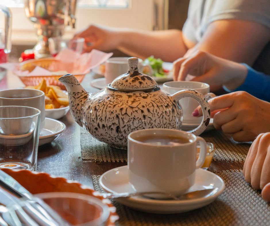Cozy morning tea at a kitchen table with a speckled ceramic teapot center stage, a steaming teacup on a saucer, bowls of snacks and hands reaching for cups.