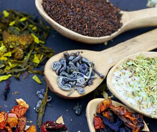 Close-up of wooden spoons holding colorful loose-leaf teas, herbal blends and dried fruit on a dark surface.