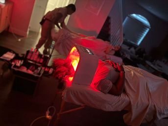 Person reclining on a spa table under a glowing red LED light therapy hood for a facial in a dim wellness treatment room, with another client and therapist in the background.