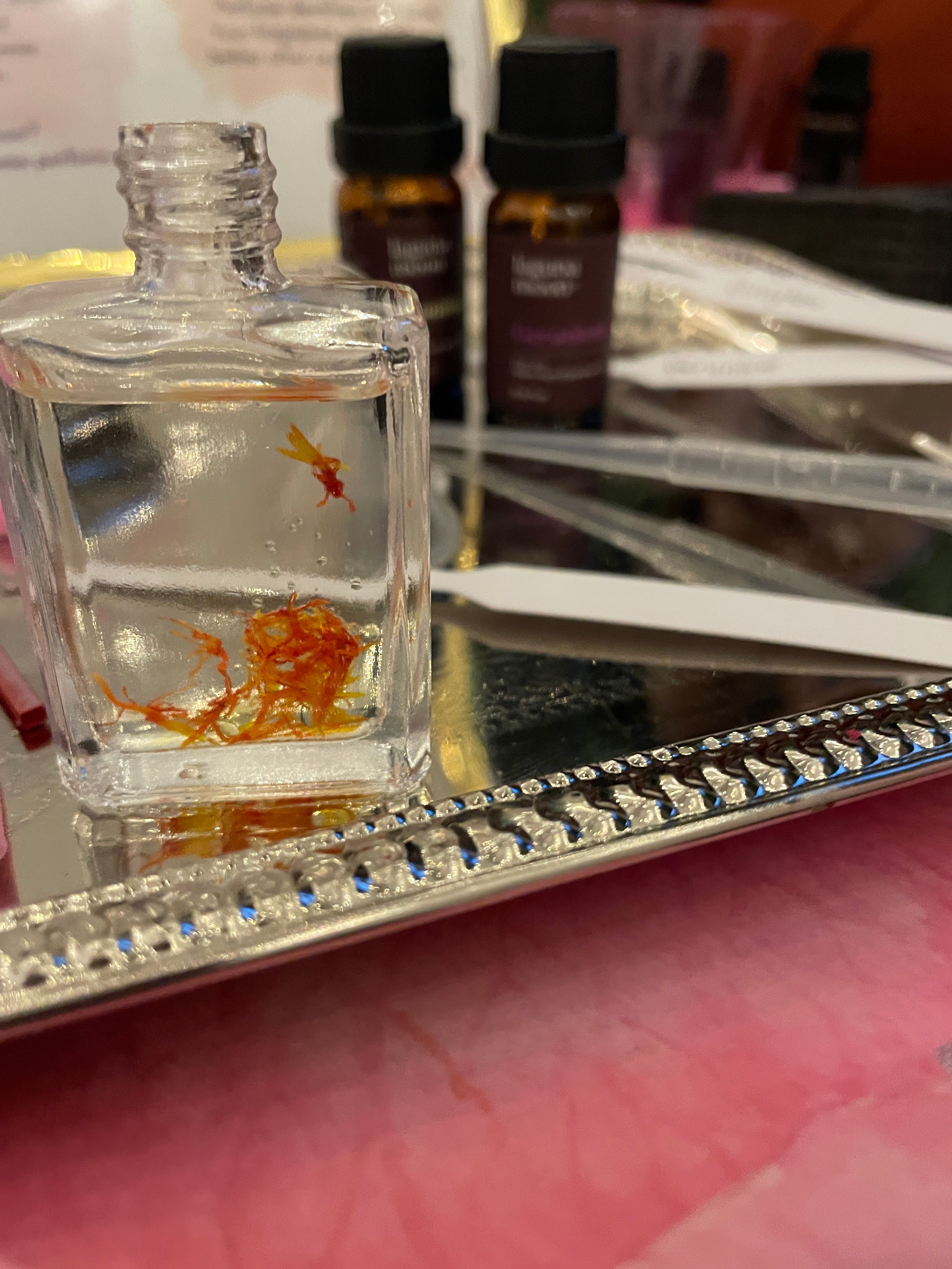 Close-up of a small clear glass bottle containing orange saffron threads on a decorative silver mirrored tray, with blurred amber essential oil vials and fragrance testing strips in the background.