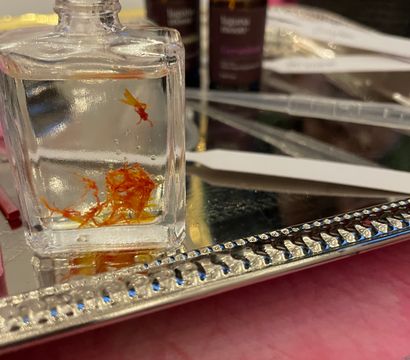 Close-up of a small clear glass bottle containing orange saffron threads on a decorative silver mirrored tray, with blurred amber essential oil vials and fragrance testing strips in the background.