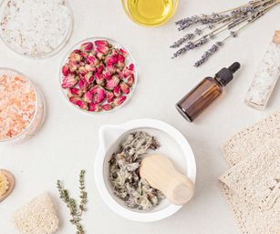 Flat lay of natural spa ingredients for DIY skincare: mortar and pestle with dried herbs, bowl of pink rosebuds, jars of bath salts, amber essential oil dropper, lavender sprigs, oil and folded towels.