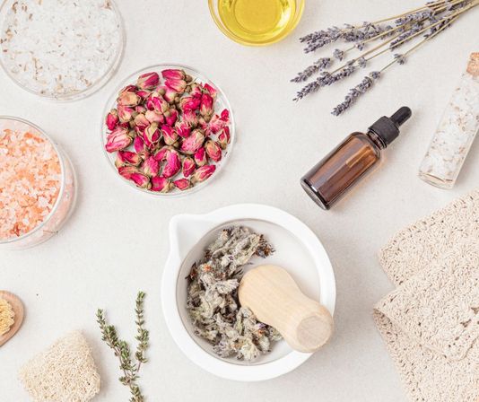 Flat lay of natural spa ingredients for DIY skincare: mortar and pestle with dried herbs, bowl of pink rosebuds, jars of bath salts, amber essential oil dropper, lavender sprigs, oil and folded towels.