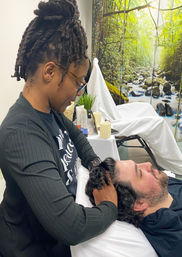 Wellness practitioner with a high dreadlock bun giving a relaxing scalp massage to a reclining client on a spa treatment table, with candles, skincare products and a soothing forest stream mural backdrop.