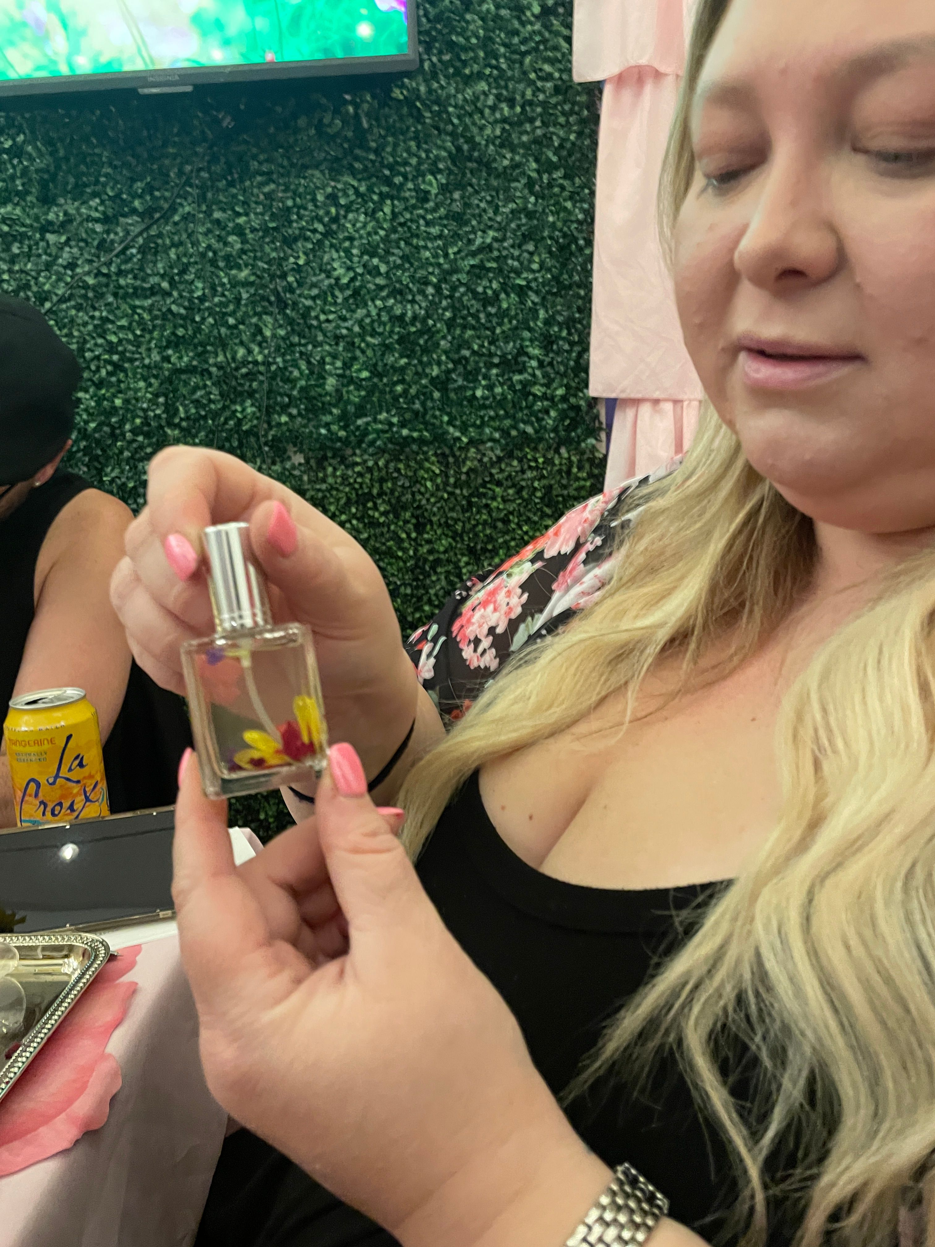 Close-up of a woman with long blonde hair holding a small glass perfume bottle filled with pressed flowers, pink nails and floral robe, seated at a decorated table with a can and green leafy backdrop.