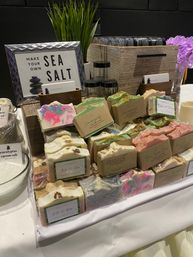 Colorful handmade artisan soap bars wrapped in kraft bands stacked on a vendor table, paired with glass jars, potted grass, purple flowers and a framed "Make Your Own Sea Salt" sign — craft market soap display.