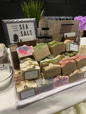 Colorful handmade artisan soap bars wrapped in kraft bands stacked on a vendor table, paired with glass jars, potted grass, purple flowers and a framed "Make Your Own Sea Salt" sign — craft market soap display.