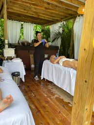 Spa therapist putting on blue gloves beside a client reclining on a treatment table in an outdoor cabana with white curtains, wooden deck and lush tropical plants — tropical resort spa setting.