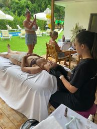 Poolside spa scene in a tropical backyard: a person reclines on a white-covered massage table while a therapist performs a head massage, with guests on wooden chairs, balloons and sun loungers beside a blue pool.